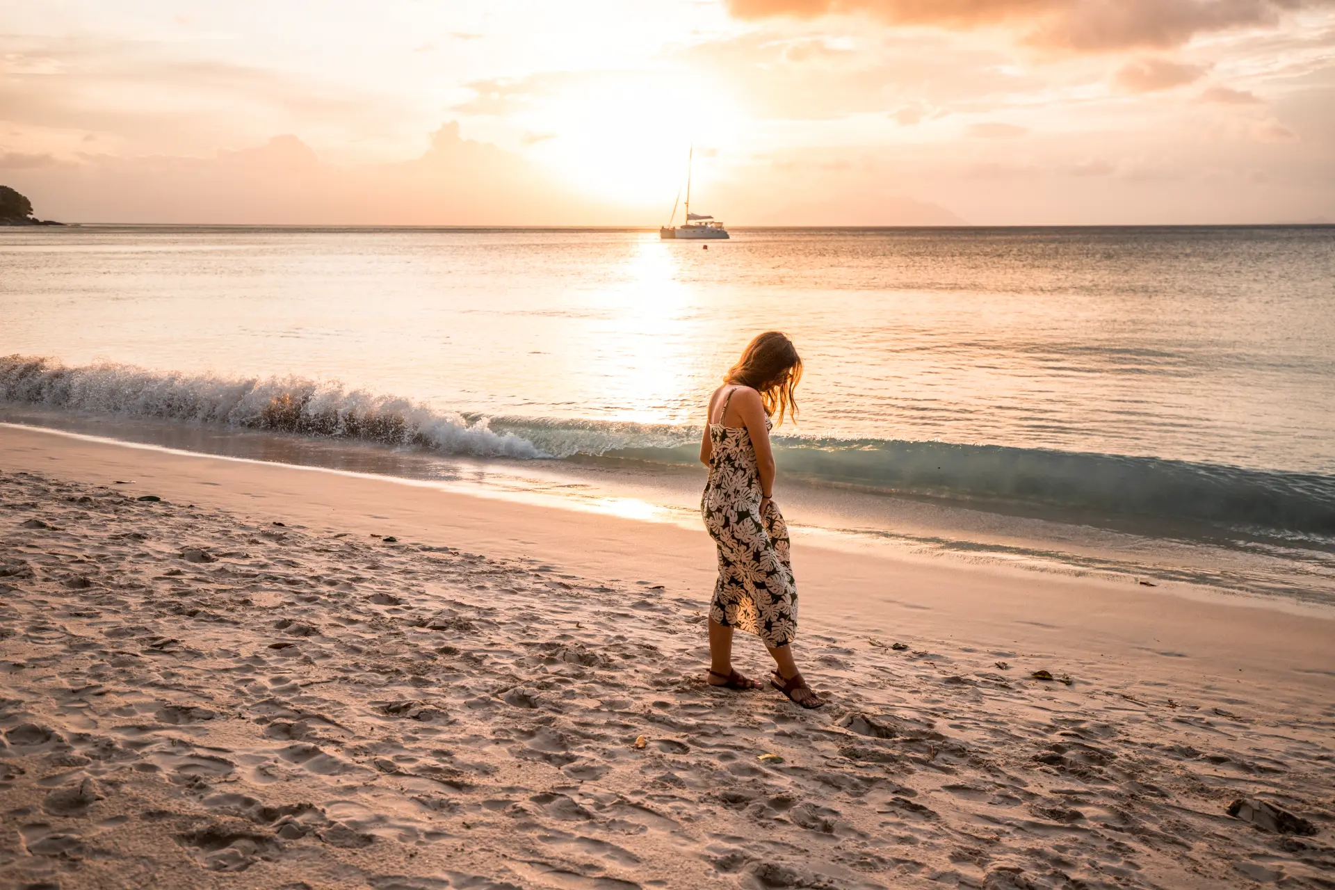 Maria che cammina sulla spiaggia al tramonto con il catamarano all'orizzonte